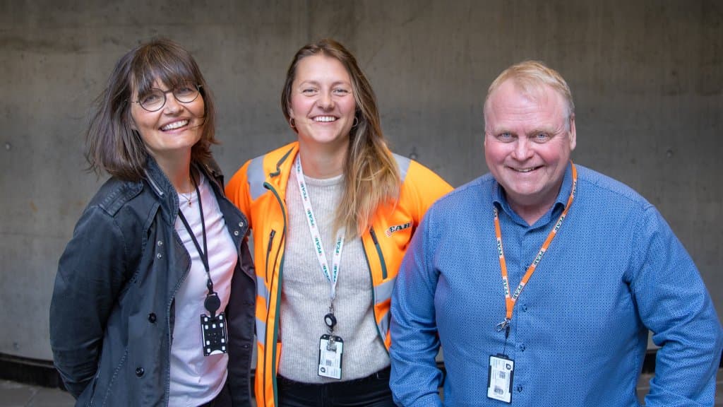 Foto av tre personer, to kvinner og én mann, som står sammen . Alle tre ser i kamera og smiler.