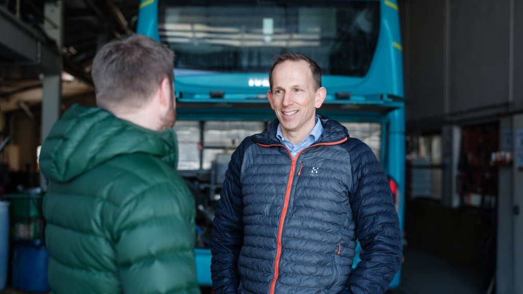 Foto av to menn som står i samtale, inne i en garasje. En av mennene står med ryggen til. Den andre mannen smiler. Han har på seg en blå jakke. I bakgrunnen er det synlig en buss.